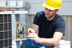 A male HVAC technician wearing a yellow hard hat and a black t-shirt, using a screwdriver to repair the electrical wiring of an outdoor air conditioning unit.
